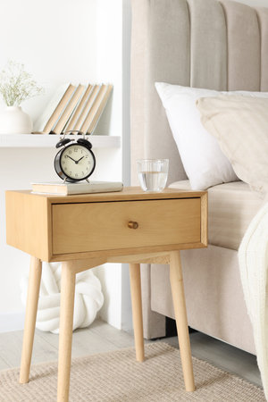 Bedside table with alarm clock, glass of water and book near bed indoors