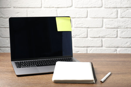 Laptop with sticky note, notebook and pen on wooden table. Space for text