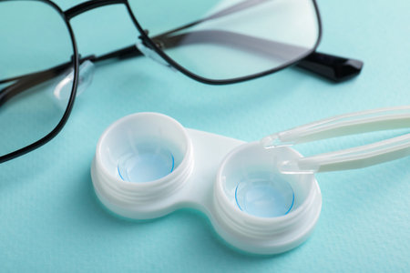 Contact lenses in case, tweezers and glasses on turquoise background, closeup