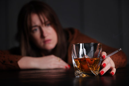 Alcohol addiction. Woman with glass of whiskey and cigarette at table indoors, selective focusの写真素材