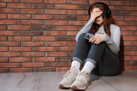 Sad teenage girl with headphones listening to music near brick wall indoors, space for textの写真素材