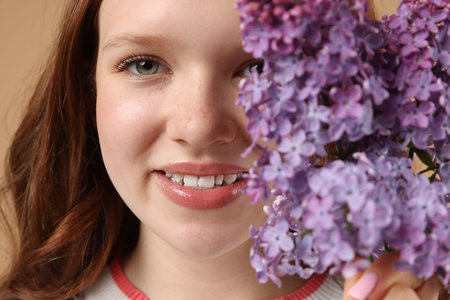 Smiling teenage girl with lilac flowers on beige background, closeup