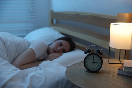Frustrated young woman suffering from insomnia on bed indoors, focus on alarm clock