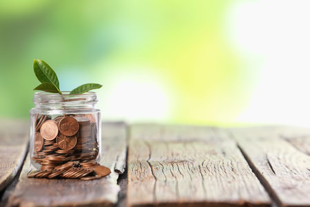 Investment concept. Glass jar with coins and sprout on wooden table against blurred green...