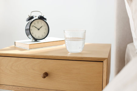 Bedside table with alarm clock, glass of water and book near bed indoors, closeup