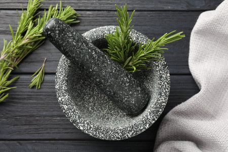 Stone mortar with rosemary and pestle on black wooden table, flat lay
