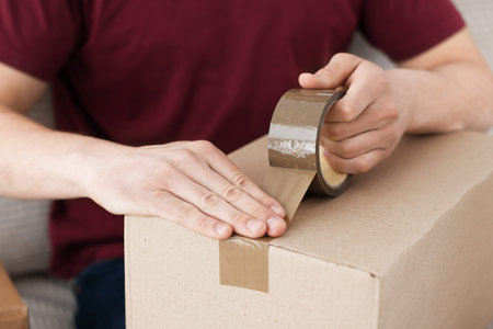 Man sealing cardboard box with adhesive tape indoors, closeup