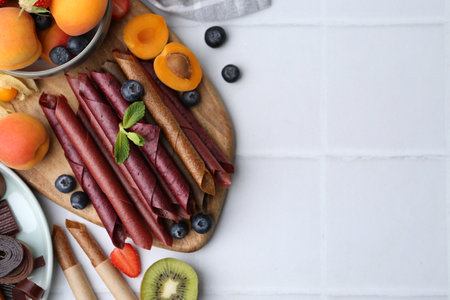 Tasty candied leather rolls, fruits and berries on white tiled table, flat lay. Space for textの写真素材