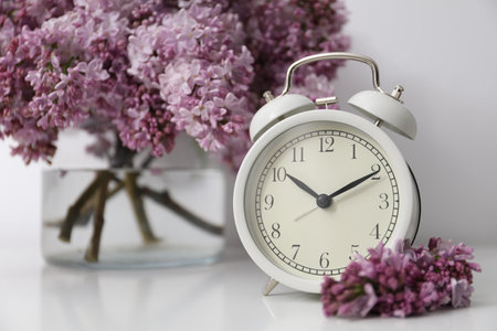 Beautiful lilac flowers and alarm clock on table near white wall indoors, closeupの写真素材