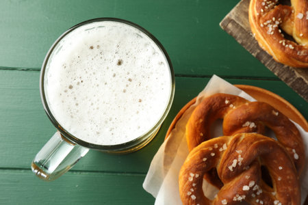 Tasty pretzels and glass of beer on green wooden table, flat lay