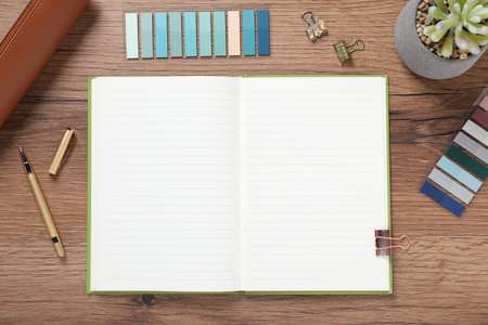 Pen, open notebook, colorful stickers, binder clips and floral decor on wooden table, flat lay