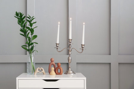 Elegant candlestick with burning candles and decor on chest of drawers near light wallの写真素材