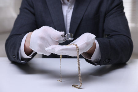 Appraiser evaluating luxury necklace at white table indoors, closeup
