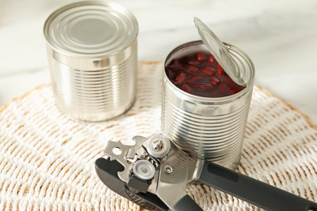 Can opener and tins with beans on white table, closeup
