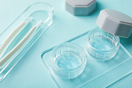 Contact lenses in case and tweezers on light turquoise background, closeup