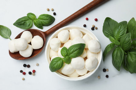 Tasty mozzarella cheese balls in bowl, spoon, basil and peppercorns on white marble table, flat layの写真素材