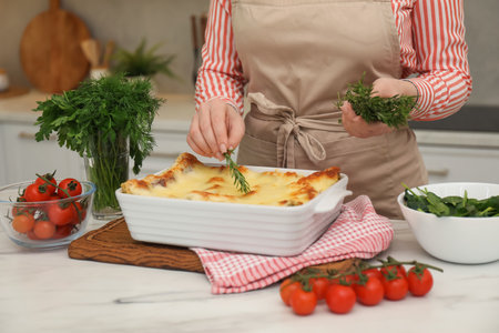Woman putting rosemary onto lasagna at white marble table in kitchen, closeupの写真素材