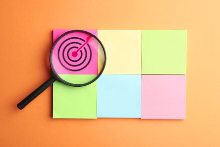 Targeting. Paper notes with bullseye and magnifying glass on orange background, top view