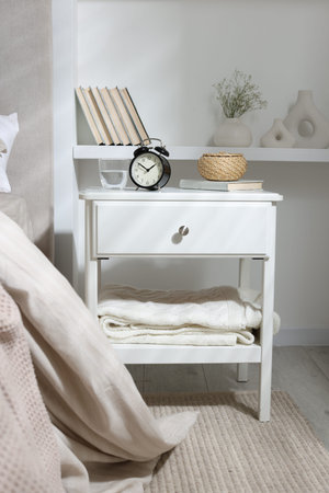 Bedside table with alarm clock, glass of water, wicker box and book near bed indoors