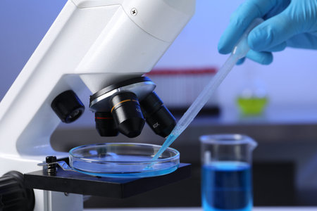 Scientist dripping liquid with pipette onto Petri dish on microscope in laboratory, closeup