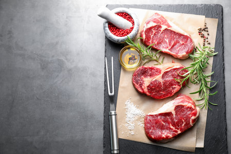 Pieces of raw beef meat, spices and carving fork on grey table, flat lay. Space for textの写真素材