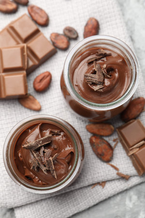 Tasty pudding in glass jars, pieces of chocolate and cocoa beans on light grey table, flat lay
