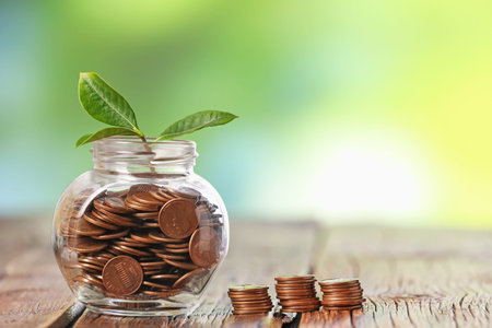 Investment concept. Glass jar with coins and sprout on wooden table against blurred green...
