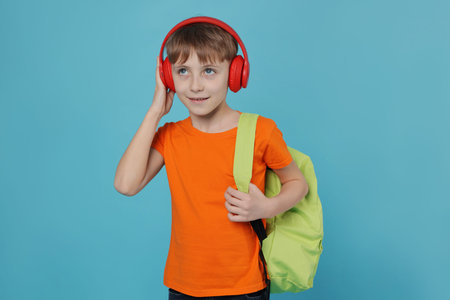 Cute schoolboy in headphones with backpack on light blue background