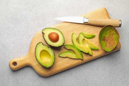 Cut ripe green avocados and knife on light grey table, top view