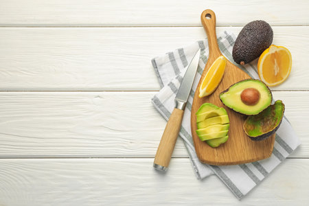 Tasty cut avocados, lemon and knife on white wooden table, flat lay. Space for text