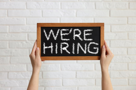 Woman holding small blackboard with phrase We Are Hiring against white brick wall, closeup. Recruitment, human resources conceptの写真素材