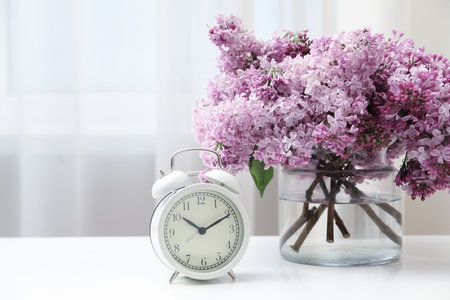 Beautiful lilac flowers in glass vase and alarm clock on table indoorsの写真素材