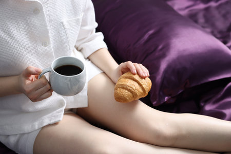 Woman with coffee and croissant on bed with purple silk linens, closeupの写真素材