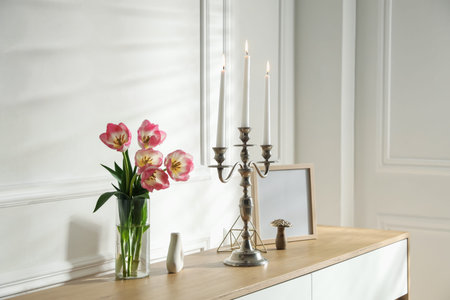 Elegant candlestick with burning candles and decor on chest of drawers near white wallの写真素材