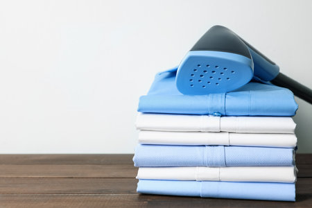 Upright steamer and stack of folded shirts on wooden table near white wall indoors, closeup. Space for textの写真素材