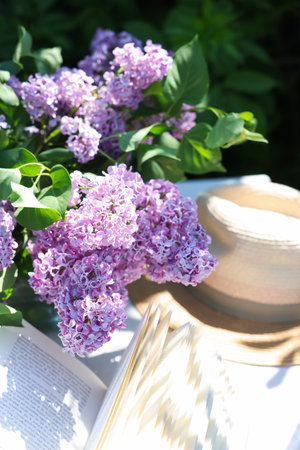 Beautiful lilac flowers, book and straw hat on table in garden, closeupの写真素材