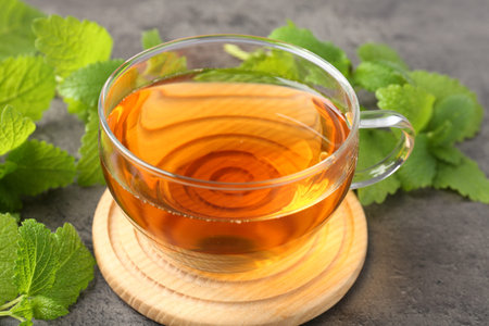 Aromatic lemon balm tea in glass cup and fresh leaves on gray table, closeup