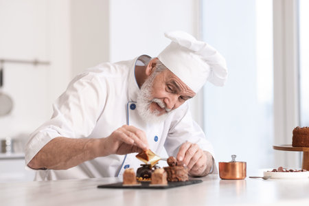Elderly pastry chef decorating tasty desserts with edible gold leaf sheet at table in kitchenの写真素材