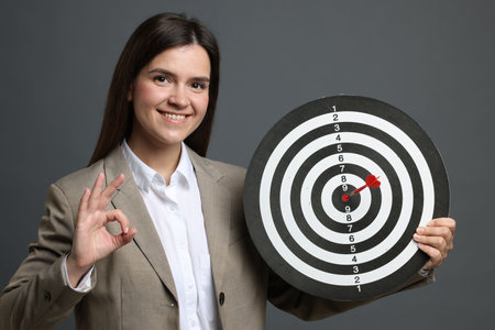 Targeting. Smiling businesswoman with dart board and arrow showing ok gesture on grey backgroundの写真素材