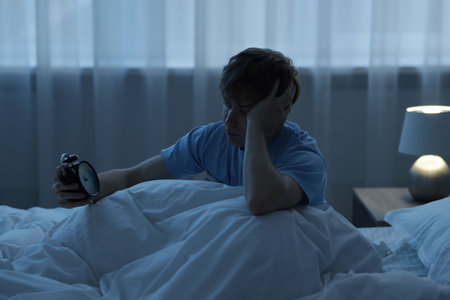 Frustrated man looking at alarm clock on bed at night. Insomnia problem