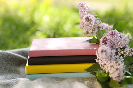 Stacked books, lilac flowers and blanket on green grass outdoors, closeup
