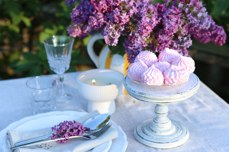 Beautiful lilac flowers, zephyrs, plates, cutlery, glasses and burning candle on table in garden