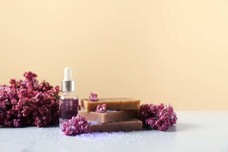 Stack of soap bars, sea salt, essential oil and lilac flowers on white table against beige background, closeup. Space for textの写真素材