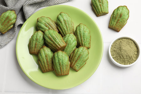 Tasty madeleine cakes and matcha powder on white tiled table, flat lay