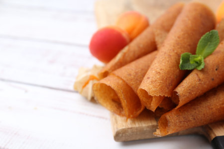 Delicious fruit leather rolls and apricots on white wooden table, closeup. Space for text
