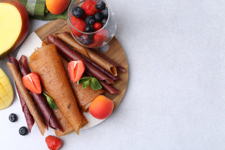 Delicious fruit leather rolls, mint, mango, apricots and berries on light table, flat lay. Space for...