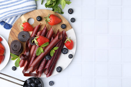 Tasty fruit leather rolls, strawberries, blueberries and mint on white tiled table, flat lay. Space...