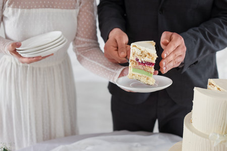 Groom putting piece of wedding cake onto plate which holding bride at table indoors, closeup
