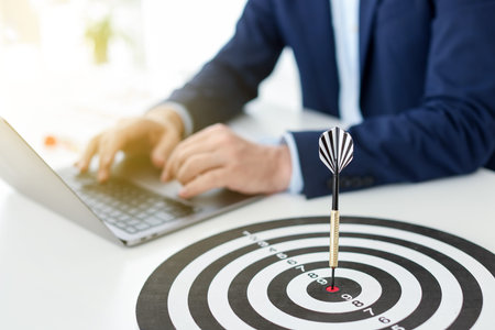 Man working on laptop at table, focus on dartboard with arrow indoors