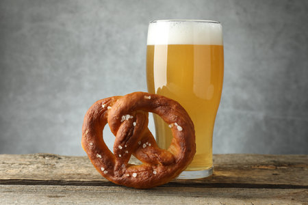Tasty pretzel and glass of beer on wooden table against grey background, closeup